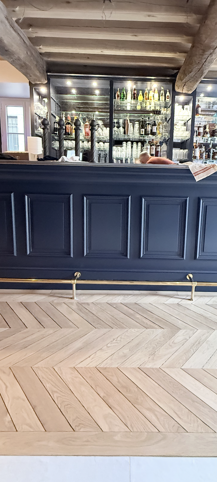 Aménagement intérieur d’un bar en pierre et bois avec parquet en chevrons, soubassement mouluré foncé et assises velours vert, signé JBB Architecture